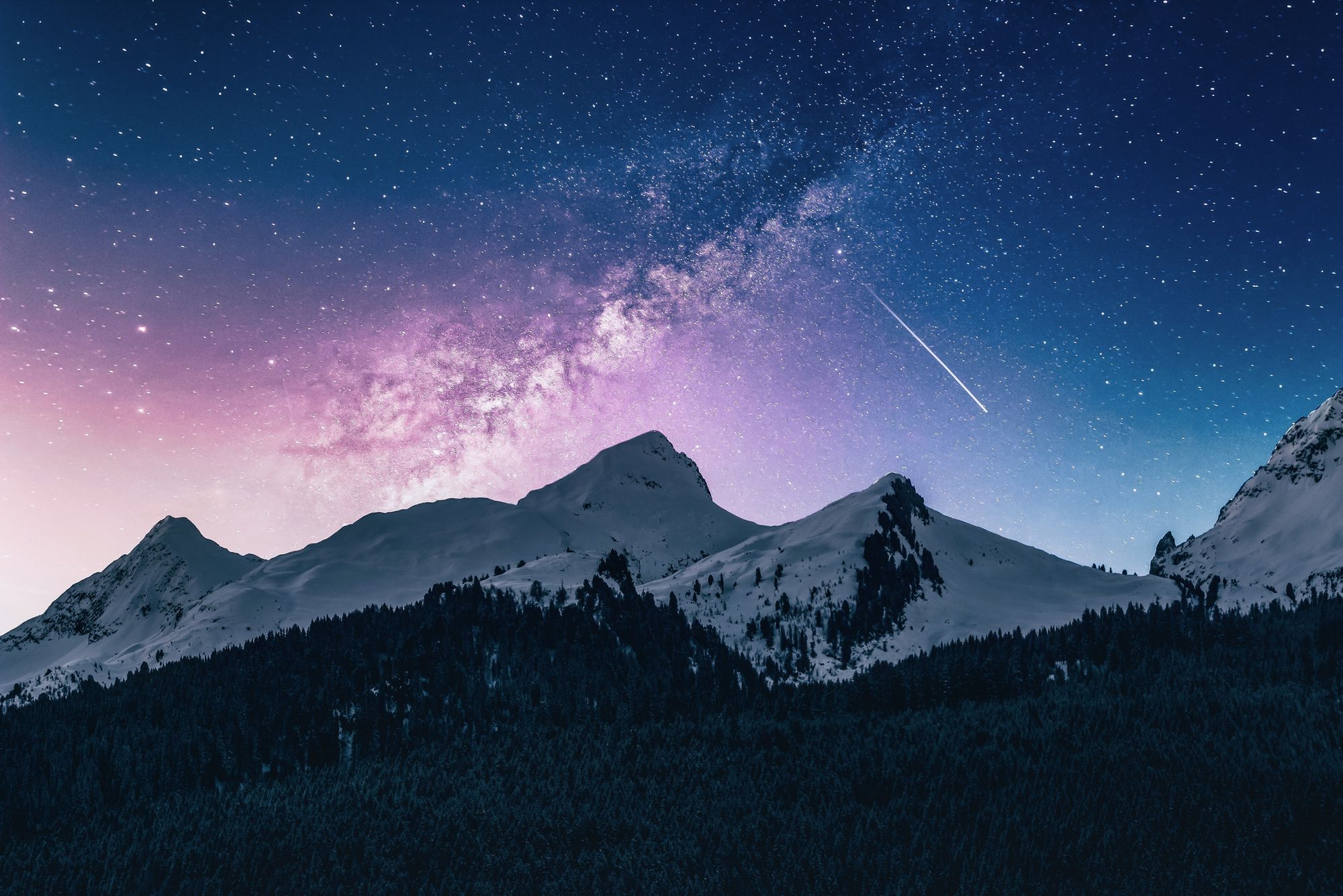 Snow-capped mountains at dusk. In the foreground, a dark forest covers the lower elevations. The sky is blue and purple with the Milky Way, numerous stars and a streaking meteor visible.