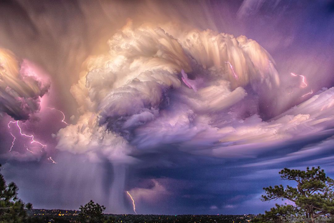 In swirling purple, blue, gray, white and orange, an anvil-shaped thundercloud hovers above Colorado Springs. Purple and gold lightning is visible on the left, in the center and up in the cloud on the right as rain comes down toward the bottom left corner. A few pine trees are visible at the bottom as are the lights of the city.