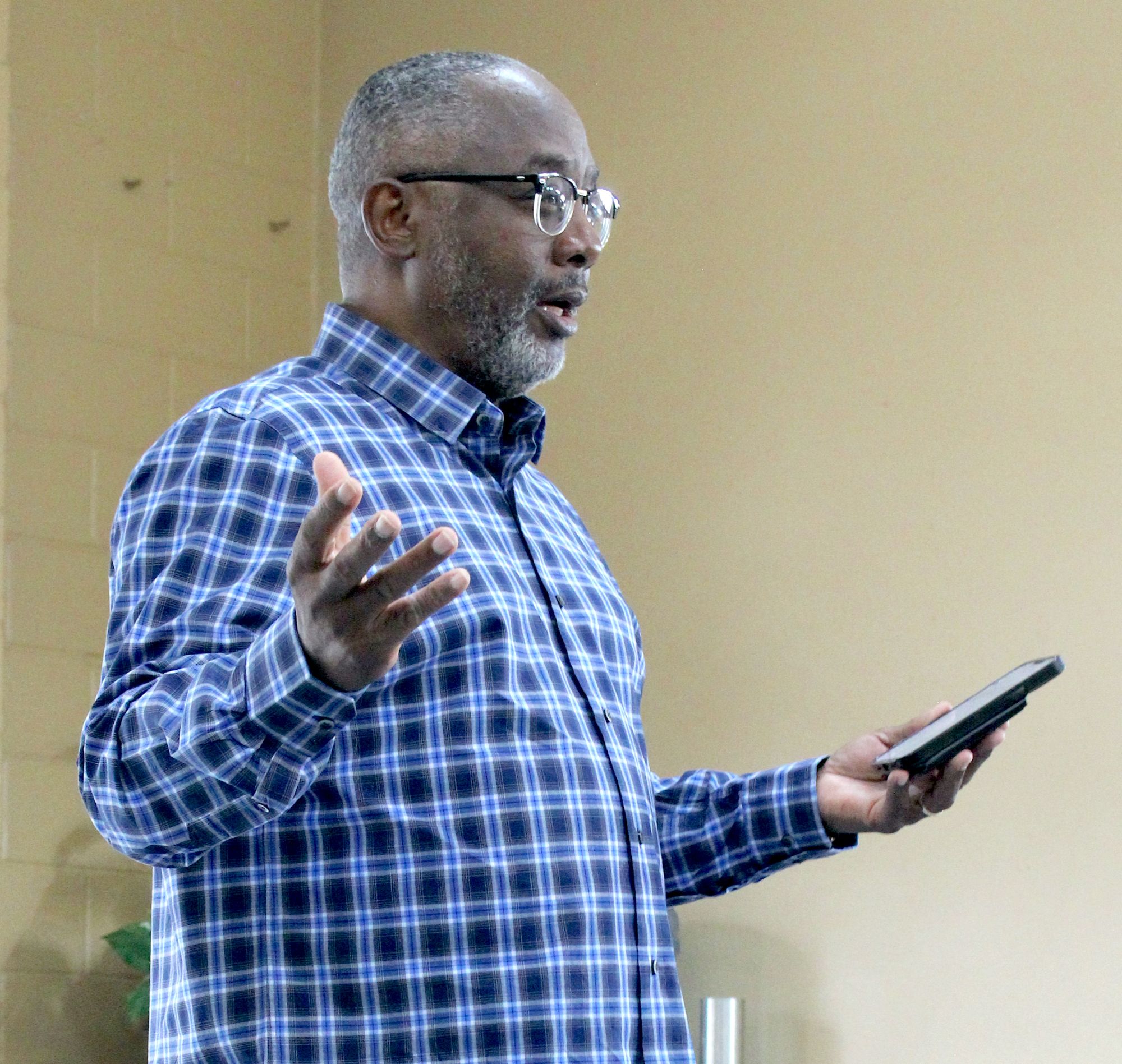 The photo is of a Black man with short, graying hair and glasses who is gesturing while holding his phone. He is wearing a long-sleeved, blue plaid shirt.