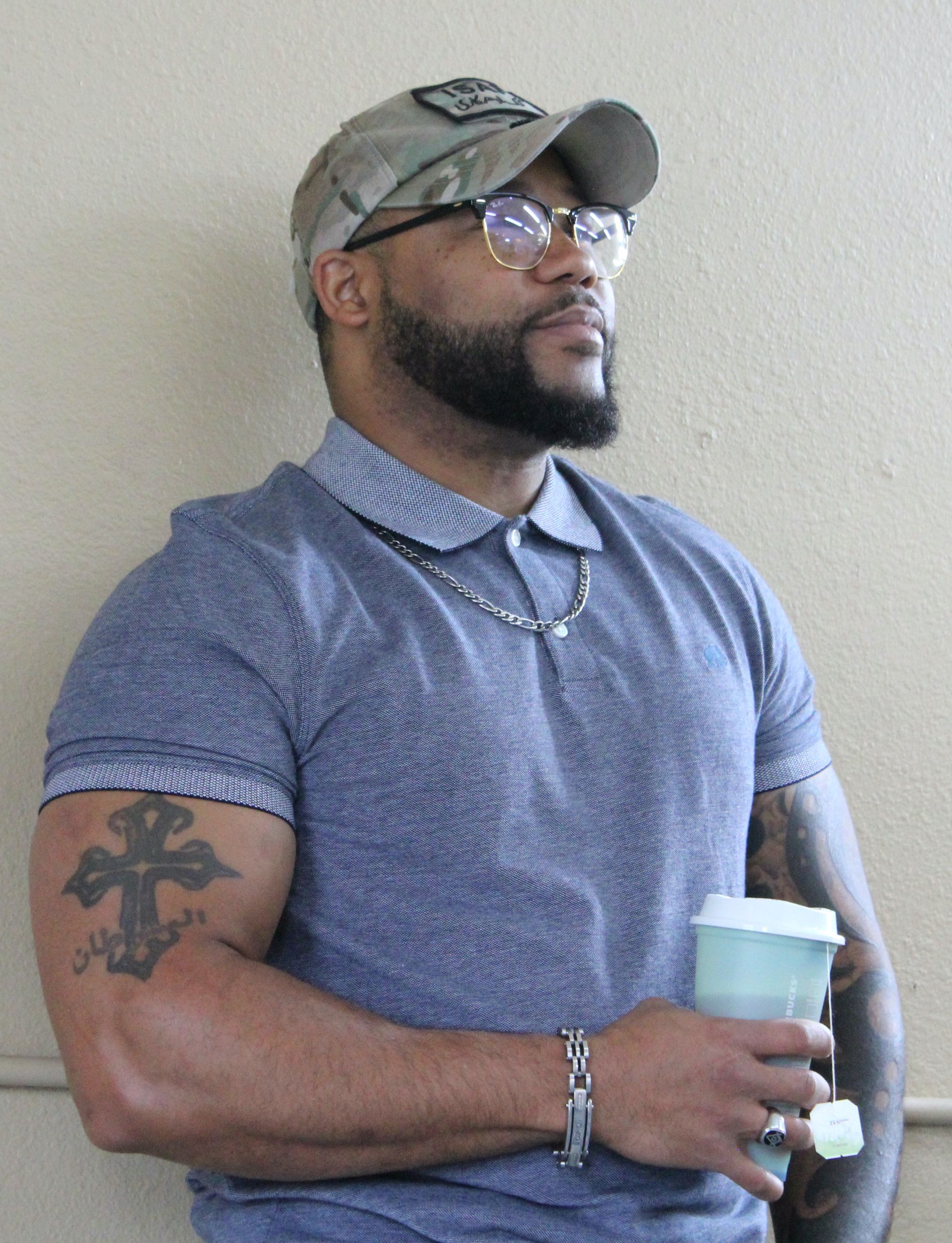 Photo of a Black man with a black beard and glasses, wearing a camouflage ball cap, a silver chain necklace and bracelet and holding a to-go cup of tea. A black tattoo of a cross is on his right arm.