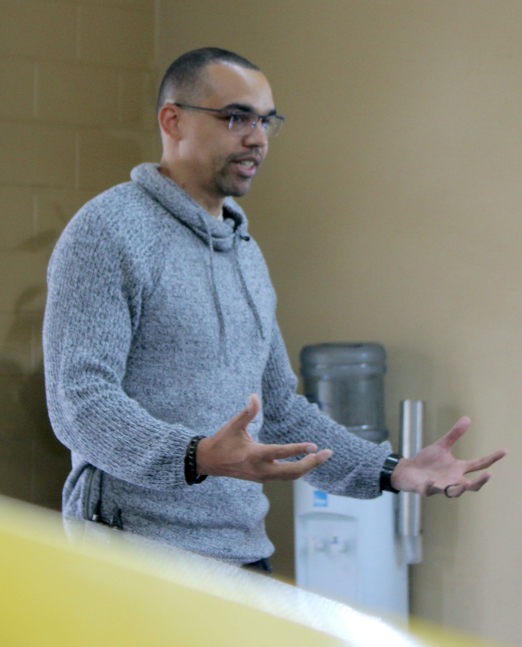 Photo of a Black man with close-cropped, black hair and five o'clock shadow. He wears glasses and a gray and white knit hoodie.