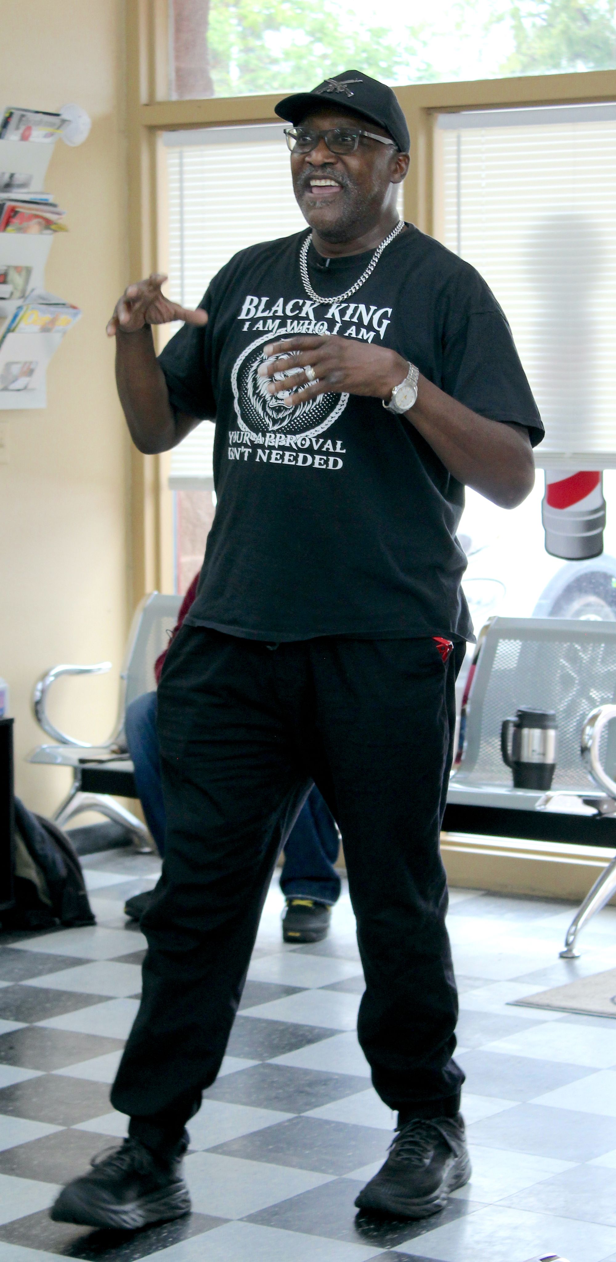 Photo is of a tall, Black man with a graying mustache and glasses. He wears a black t-shirt, silver chain and silver wristwatch.