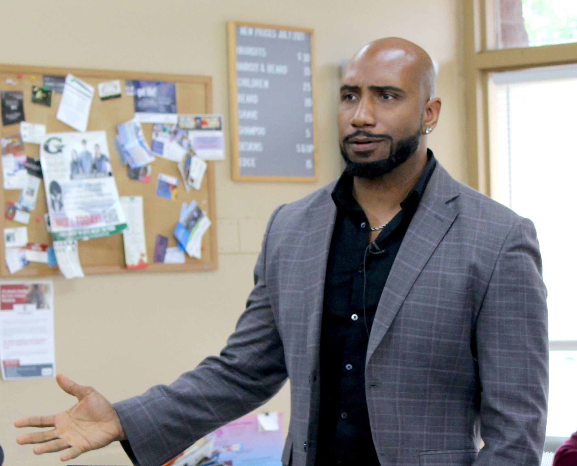 Photo of a bald Black man with a cropped, black beard. He is wearing a black dress shirt and gray suit jacket, a silver chain and diamond studs in his ears. 