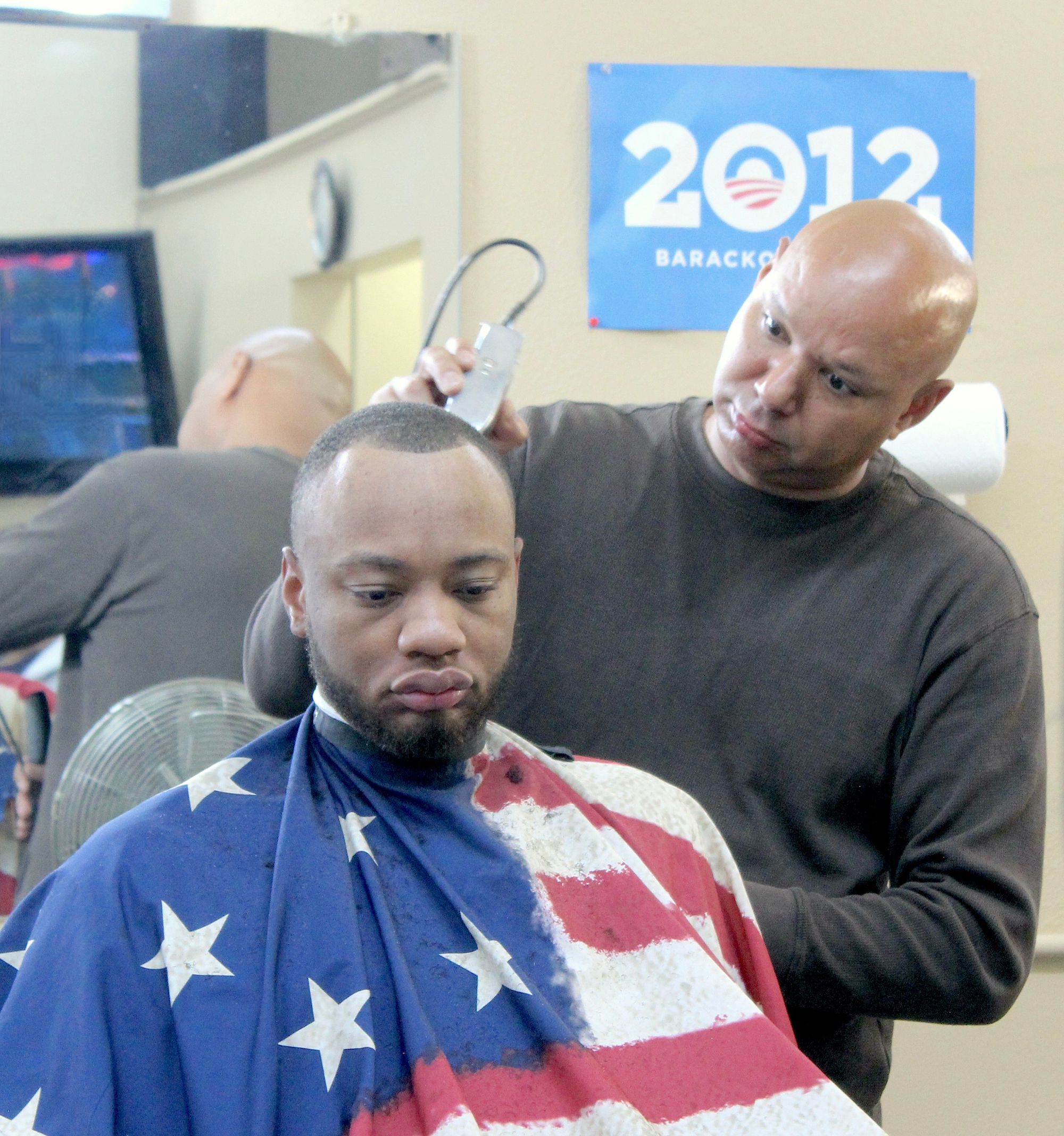 Photo of a barber trimming a man's hair. The barber is bald and the customer is wearing a drape with an image of the American flag on it. In the background, their reflection is visible in the mirror as is a small blue, white and red poster for Barack Obama's campaign in 2012.