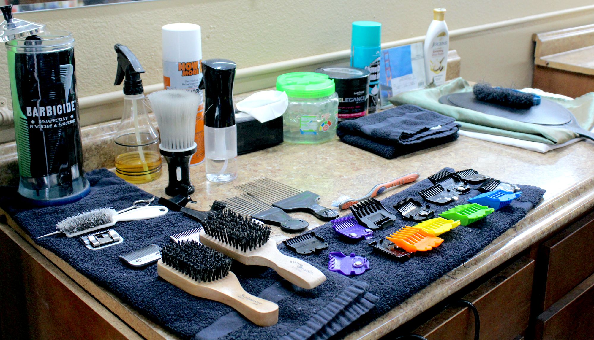 A variety of barber tools are organized neatly on two navy blue towels, including a tall jar of Barbicide with combs inside, 2 hairbrushes, a whisk-type brush, several jars and spray containers and 2 rows of trimming attachments in purple, orange, green, yellow, blue and black.