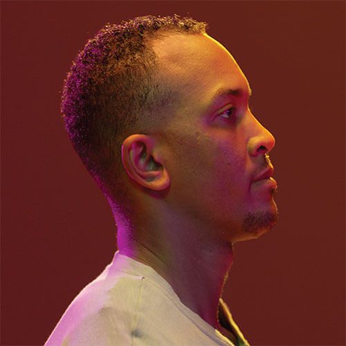 Profile photo of a young, Black man with short, curly hair and light stubble. He faces rights. He wears a pale t-shirt.