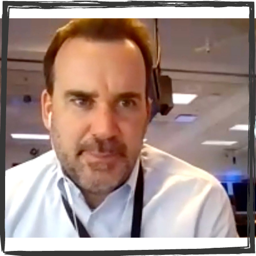 Photo of a white man w/brown hair & brown, stubbly facial hair wearing a white, collared shirt