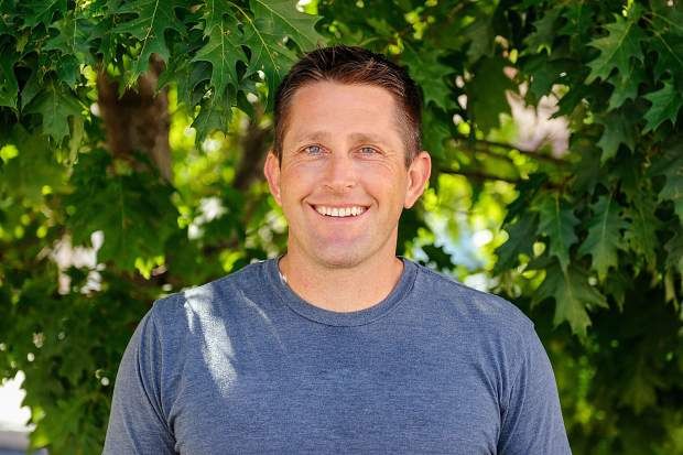 Photo of a white man with short brown hair, smiling, wearing a blue sweater & standing in front of an oak tree