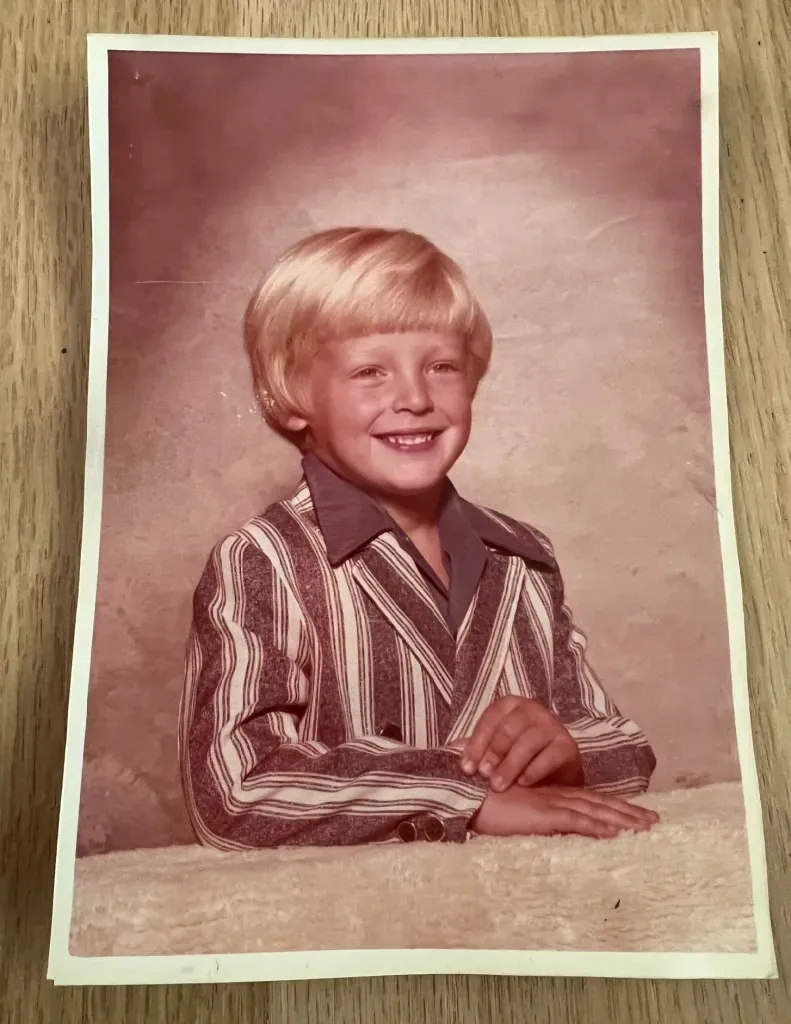 A young boy with short, blond hair smiles at the camera. he wears a brown-and-white striped suit jacket and brown shirt w/long collar. His hands are crossed in front of him on a counter.