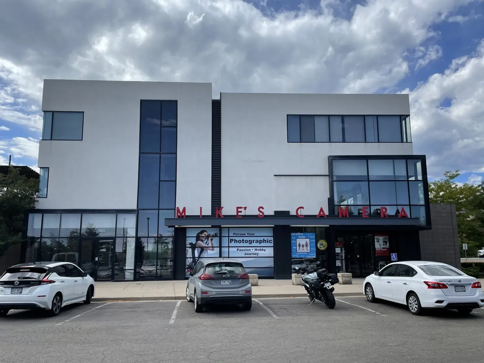 A contemporary, white, 3-story building with large windows framed in black, the store name, "Mike's Camera," and individually placed, red letters on the awning in front of the store. Three cars and a motorcycle are parked in spaces in front of the store.