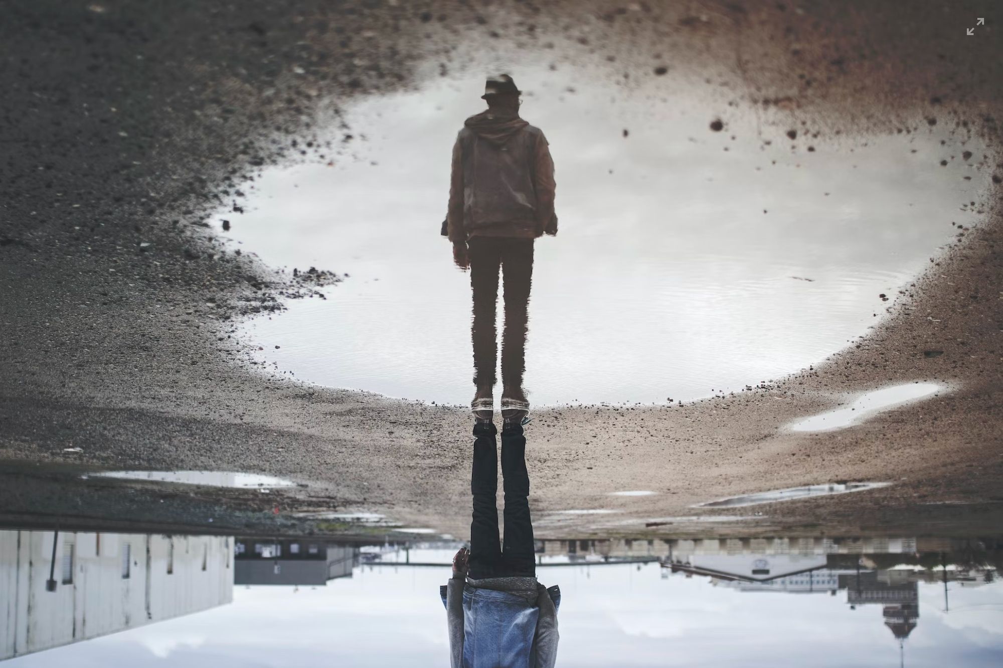 Photo of a man's reflection in a puddle. The reflection shows he's wearing a hat and coat. He stands near a warehouse