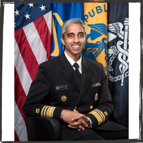 An man of Indian ethnicity wears a black uniform ew/gold braids in the sleeves and ribbons. In back are the US flag & 3 others