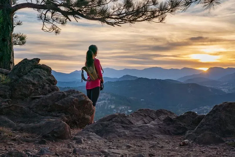 A woman w/long, dark hair in a ponytail and wearing a red shirt, leggings and a backpack looks toward the sunset from a mountain outcrop