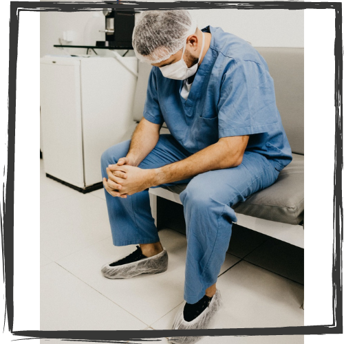 A male nurse in scrubs, face mask and shoe covers sits on a chair with hands clenched staring at the floor