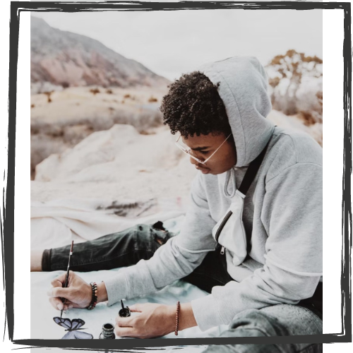 A Black teen wearing glasses & a gray hoodie sits on the ground w/a bottle of ink and pen drawing a butterfly