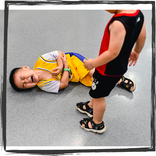 A small, Asian-looking boy lies on a floor, clutching himself and crying as another boy looks on