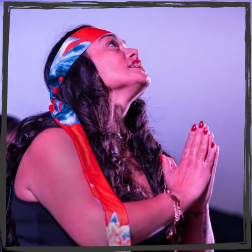A Latina woman w/long, dark hair and wearing a colorful, red head scarf prays and looks up