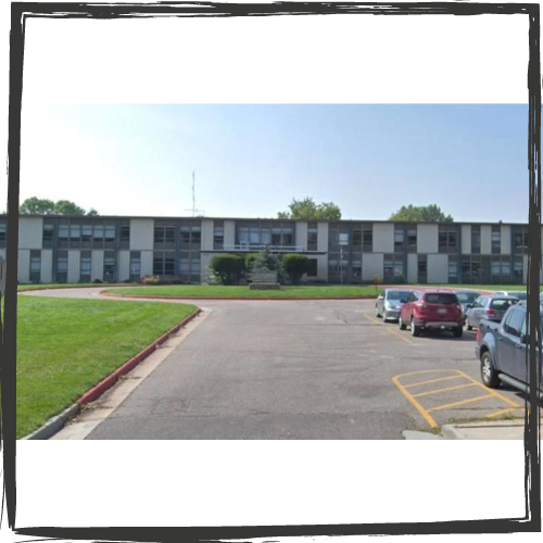 A 2-story building with a white facade pierced by multiple windows. A parking lot with several cars is in front