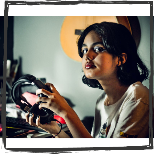 A girl w/olive skin and shoulder-length, black hair holds headphones and looks stoically at the viewer