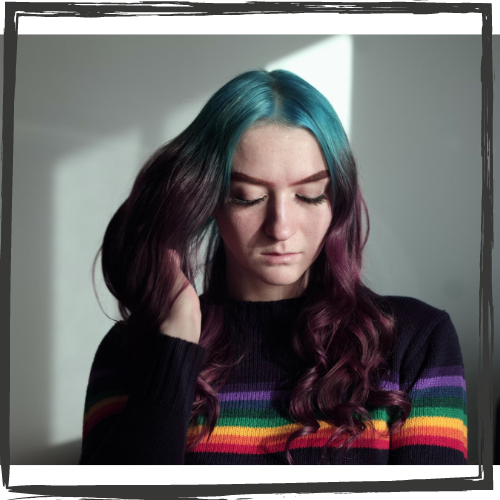 A young, white girl w/shoulder-length blue & purple hair wearing a sweater w/rainbow stripes