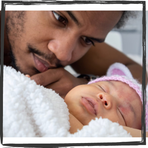 A young, Black man w/a mustache gazes at his sleeping newborn wearing a pink, knit cap