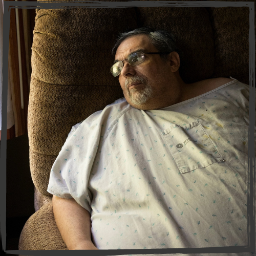 An obese man w/short, iron gray hair & wearing glasses sits in a chair