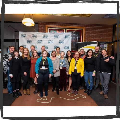 17 people stand as a group in front of a "This Is My Brave" background and smile in the theater lobby