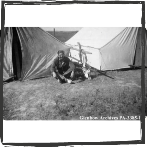 A b&w photo of Maslow sitting on the ground cross-legged in front of two canvas tents