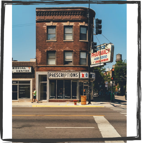 An old 3-story, brick building on a corner contains a pharmacy on the 1st floor; a sign for it hangs out front