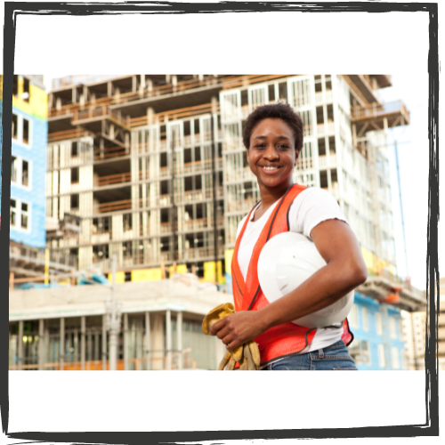 A black woman construction worker stands in front of a tall building under construction