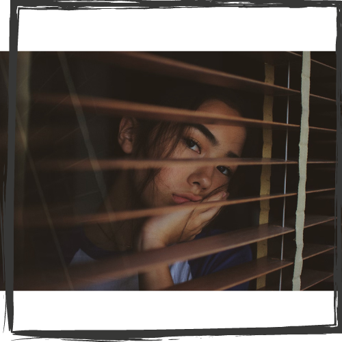 A young woman w/dark hair and eyebrows appears sad as she looks out between wood blinds