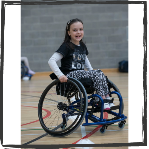 A girl w/long, dark hair wearing a long-sleeve shirt sits in a wheelchair on a basketball court