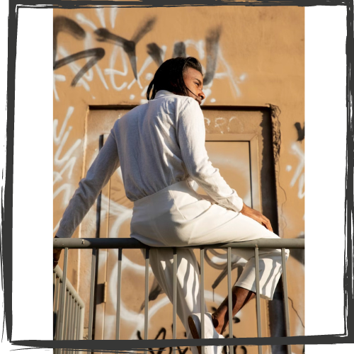 A young man w/dark, shoulder-length hair and wearing a white shirt and slacks sits on a guardrail in front of a tan graffiti wall