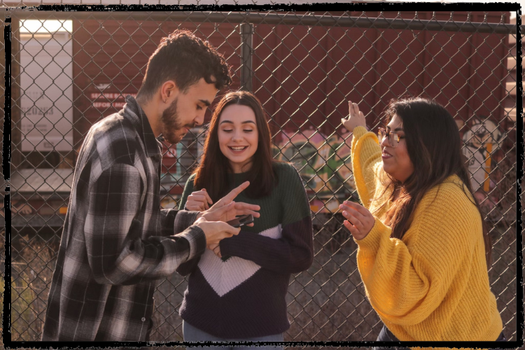 3 teens, 2 girls and 1 boy, converse in front of a chainlink fence