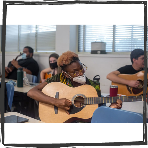 A Black woman with glasses plays a guitar while sitting as others behind her do the same