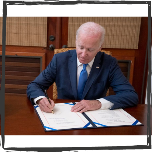 Pres. Biden sits at a wood desk signing two, large papers with blue ribbons on the corners