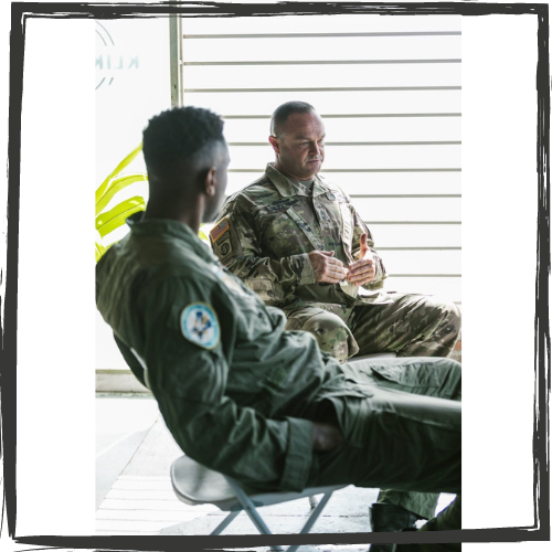 A Black man and a white man wearing military fatigues talk to each other while sitting