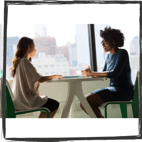 A young woman w/long hair chats at a table with a Black woman wearing glasses