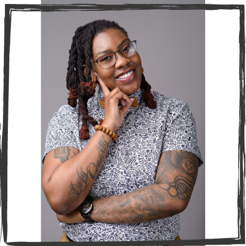 Photo of a Black queer person with shoulder length, thick braids & wearing glasses and a short-sleeved white shirt w/a black geometric design