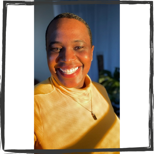 A Black woman with short, black hair smiles broadly; she wears a bright yellow top & the sun from a window shines on her face