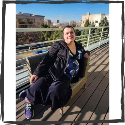 A white Transgender person poses on a bench near a railing; she has short, black hair and wears a dark knit outfit w/a blue, black & white blouse