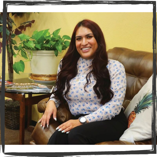 Photo of a Latina Transgender woman with long, dark hair & wearing a knit shirt w/small blue flowers; she sits on a sofa