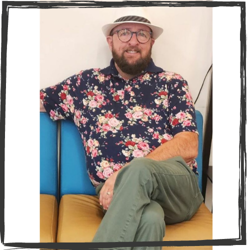 Photo of a white, cis man wearing a flowered shirt, glasses and a white hat. He sits on a sofa with 1 leg crossed over at the knee