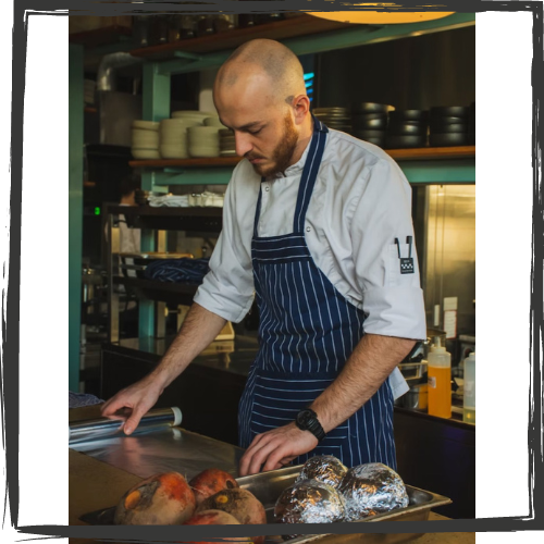 Photo of a man wearing a lue apron w/white stripes cutting up food at a restaurant counter