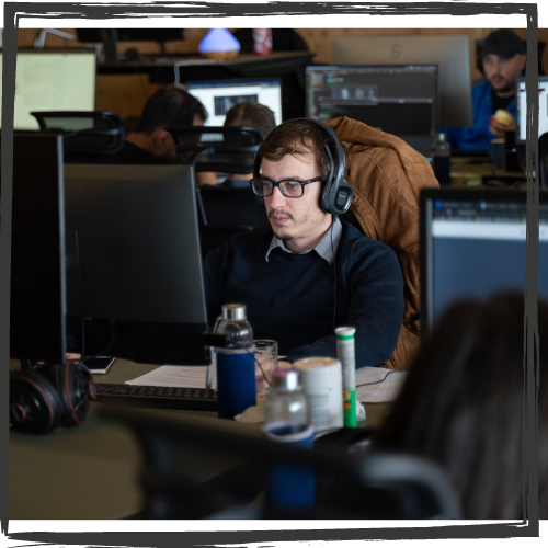 Photo of a man wearing headphones and glasses sitting at a desk & staring at a computer screen