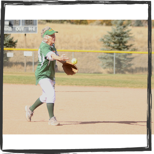 Photo of an older woman w/curly gray hair wearing a green-and-white jersey catching a softball