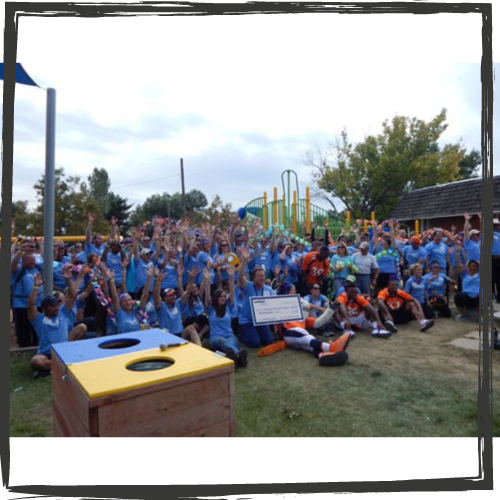 Photo of a large group of high-school aged students, all wearing blue t-shirts and raising their arms in celebration, pose standing or seated in a grassy area