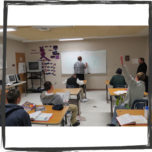 Photo of a teacher writing on a white board and high school-aged students sitting at desks; one has his arm raised; all are facing away from the camera