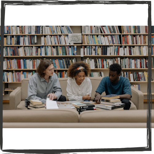 Photo of 3 diverse teenage students sitting on a sofa in a library in front of a low table covered with books