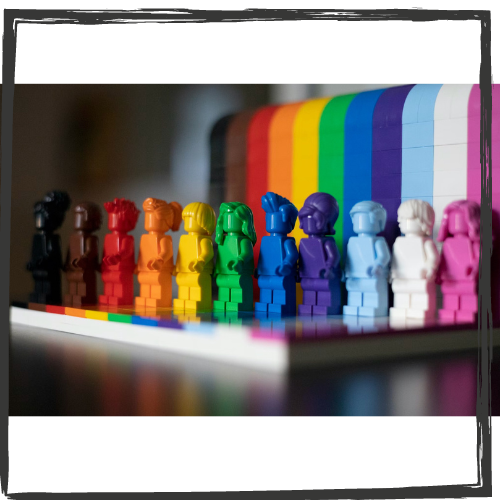 Photo of 11 toy figures of standing people, each one color of a rainbow with a rainbow cloth draped behind them